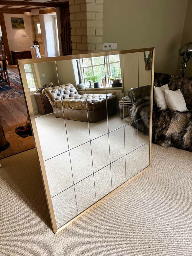 An antique mirror with a brushed brass frame rests on the carpeted floor, reflecting a cozy living room. The space features a brown sofa, a stylish armchair draped with a fur throw, and a window offering views of lush greenery outside.