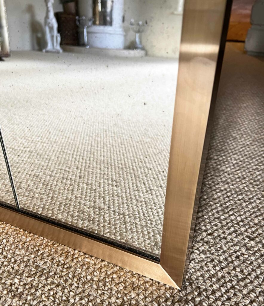 Close-up of a large antique mirror with a gold frame, angled on a textured beige carpet. The reflection reveals part of the room, including a fireplace and decorative items, slightly out of focus.