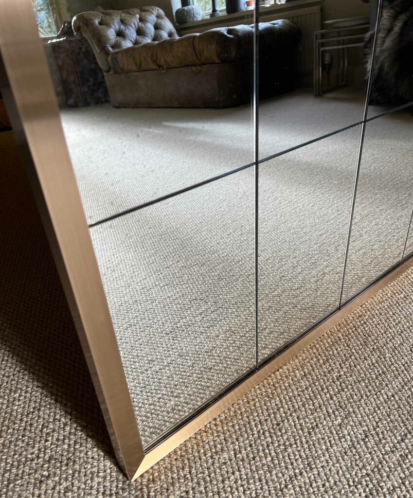 A close-up of a large bespoke mirror with a gold frame, reflecting a carpeted room with a tufted sofa and a window in the background. The image captures the texture of the carpet and the detailed reflection of the interior.