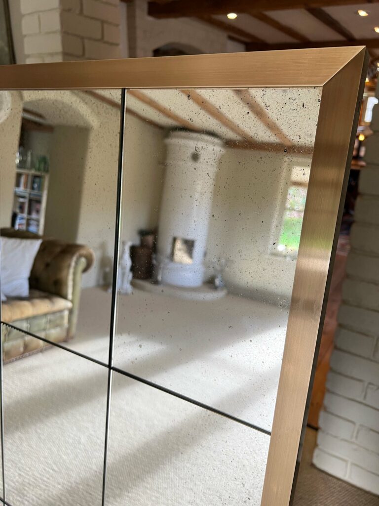 A close-up of a large bespoke mirror reflecting a cozy living room. The room features a white cylindrical fireplace and a brown leather sofa. The mirror's surface shows a grid pattern, adding a modern touch to the rustic decor.