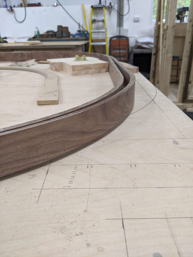 Curved wooden pieces on a workbench in a workshop. Drafting lines and measurements are visible on the table surface. Shelves, tools, and a ladder are in the background.
