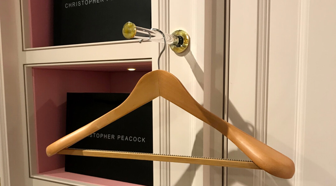 A wooden coat hanger is hanging on a brass and acrylic valet hook in front of a white wardrobe with pink shelves.