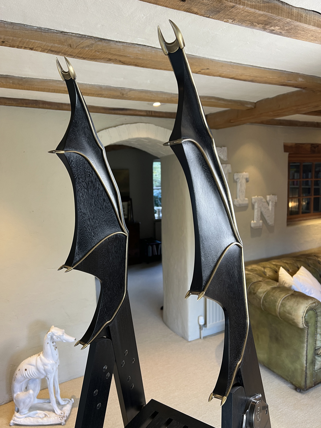A pair of ornate, black and gold chair backrests shaped like bat wings, attached to a black chair. In the background, there is a white statue of a dog, a green sofa, and wooden beams on the ceiling.