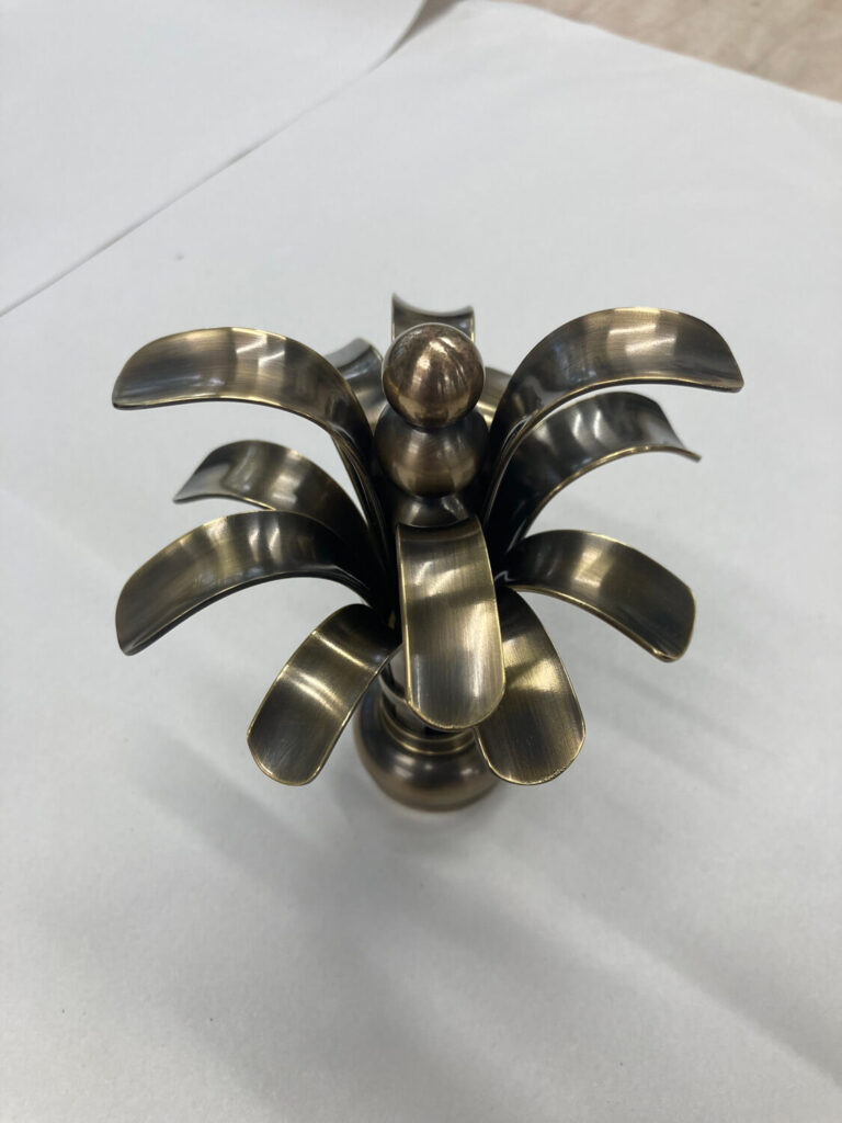 A brass decorative piece resembling a stylized palm tree with curved, petal-like leaves, placed on a white surface.