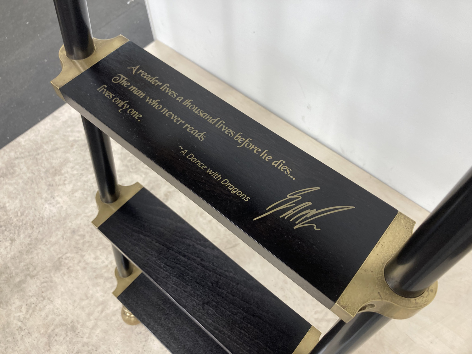 A black shelf with a gold quote that reads: A reader lives a thousand lives before he dies. The man who never reads lives only one. - A Dance with Dragons, with a gold signature underneath.