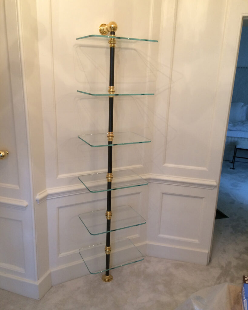 A modern corner shelving shoe rack with five glass shelves supported by a black and brass vertical pole, installed against white paneled walls in a carpeted room.