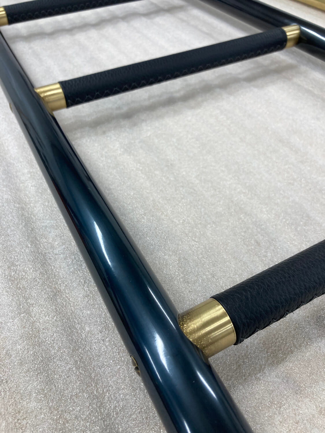 A close-up of a ladder with sleek, black side rails, brass fittings, and black leather steps, placed on a light beige carpeted surface.