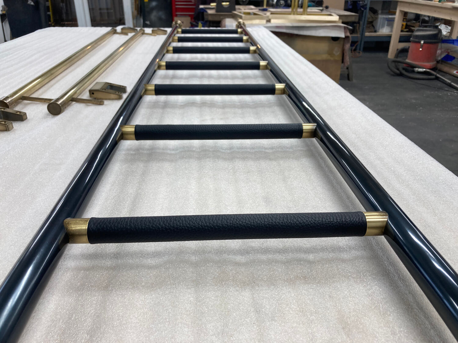 A close-up view of a metal ladder with black leather rungs and brass accents, laid out on a padded white surface in a workshop with various tools and tables in the background.