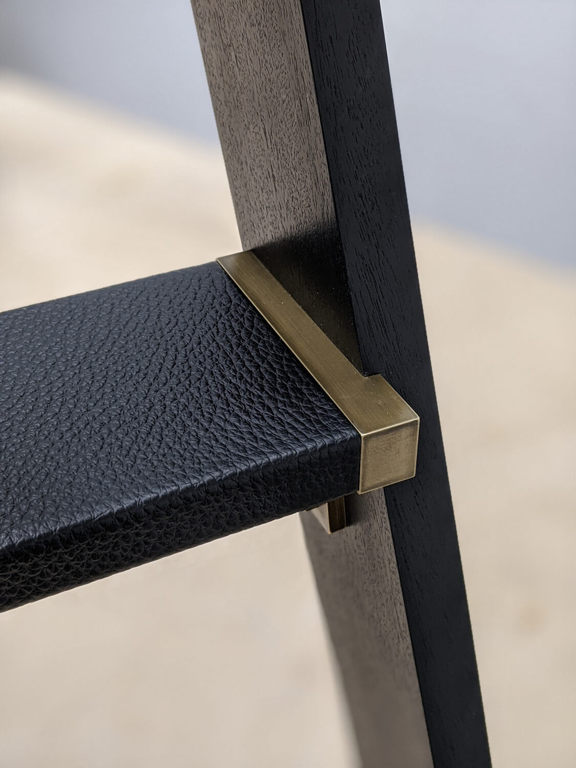 Close-up of a modern step ladder, showing the corner where a textured black leather step meets a dark wooden frame, joined with a brass metal bracket. The background is blurred in neutral tones.