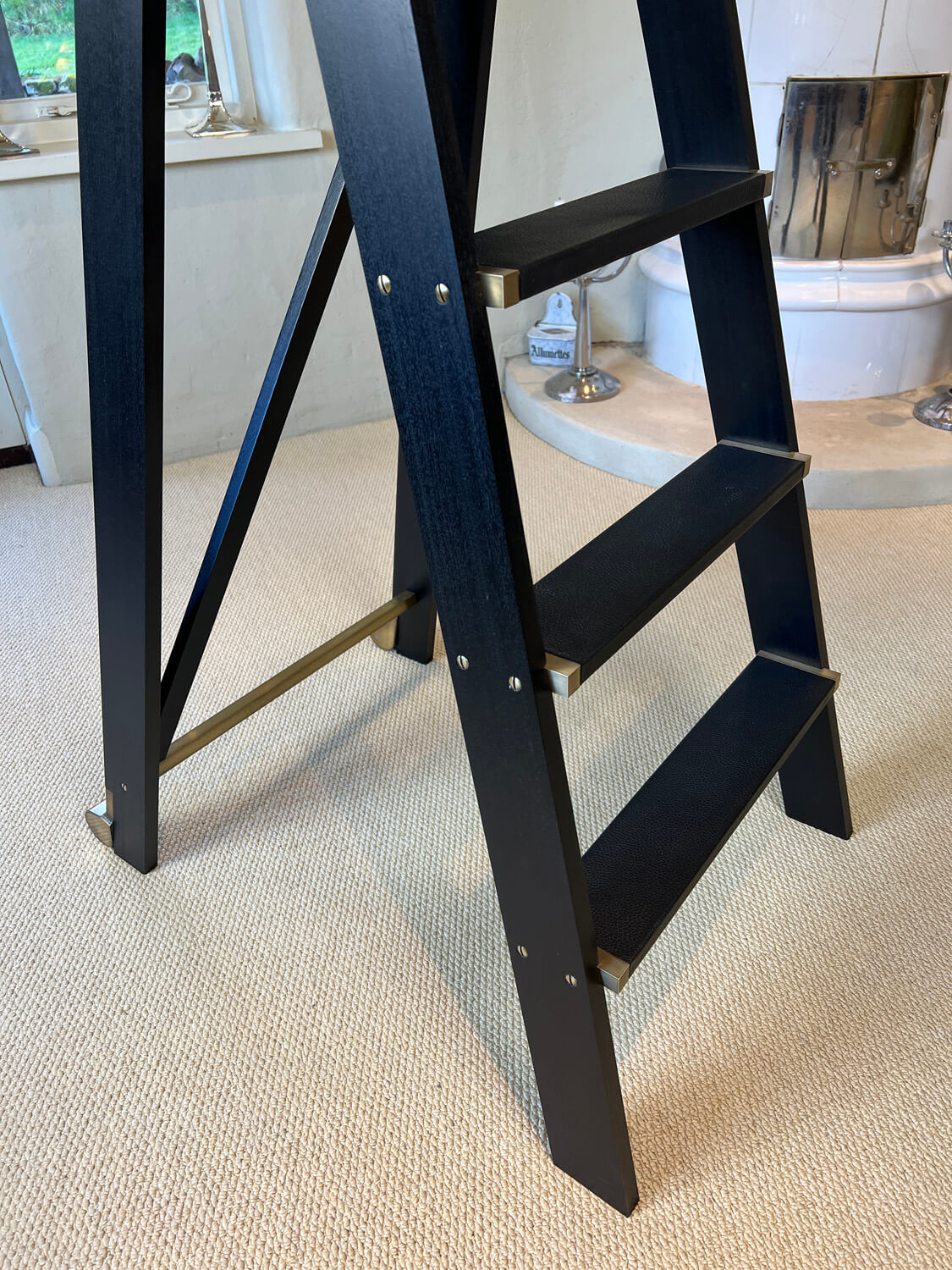 A close-up of a black wooden step ladder with brass supports standing on a beige carpet in a room with a bathtub and decorative items in the background.