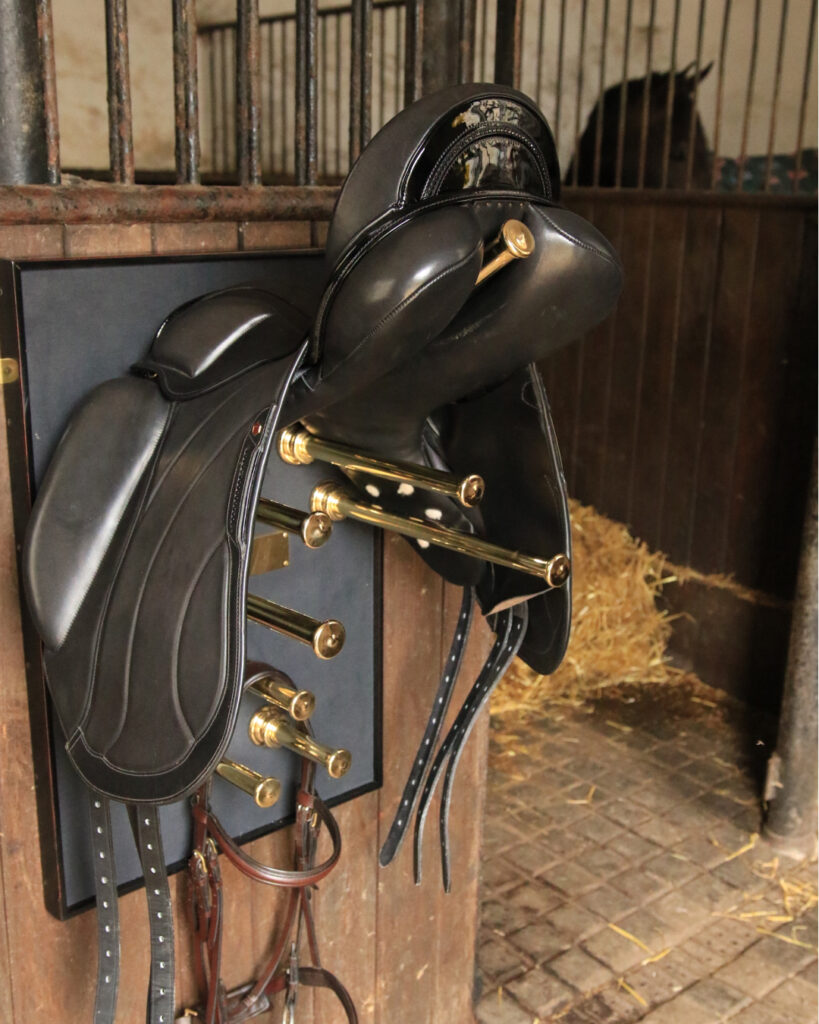A close-up of black leather saddles neatly hung on a brass saddle rack attached to a stable wall, with straw and part of a horse visible in the background.