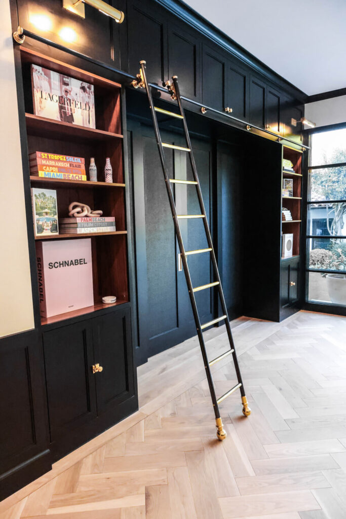 A black built-in bookshelf with brass accents features a rolling library ladder. The shelves display books and decor, while herringbone flooring and large windows fill the room with natural light.