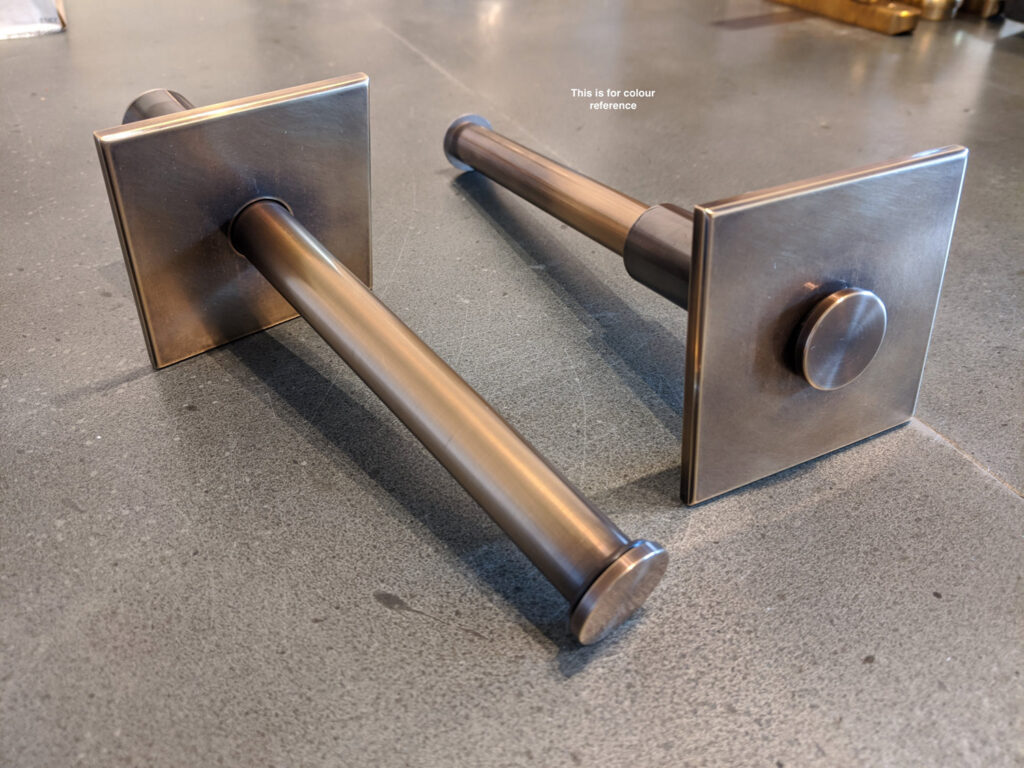 Two metallic, square plated, valet rods with a bronze finish are lying on a gray surface.