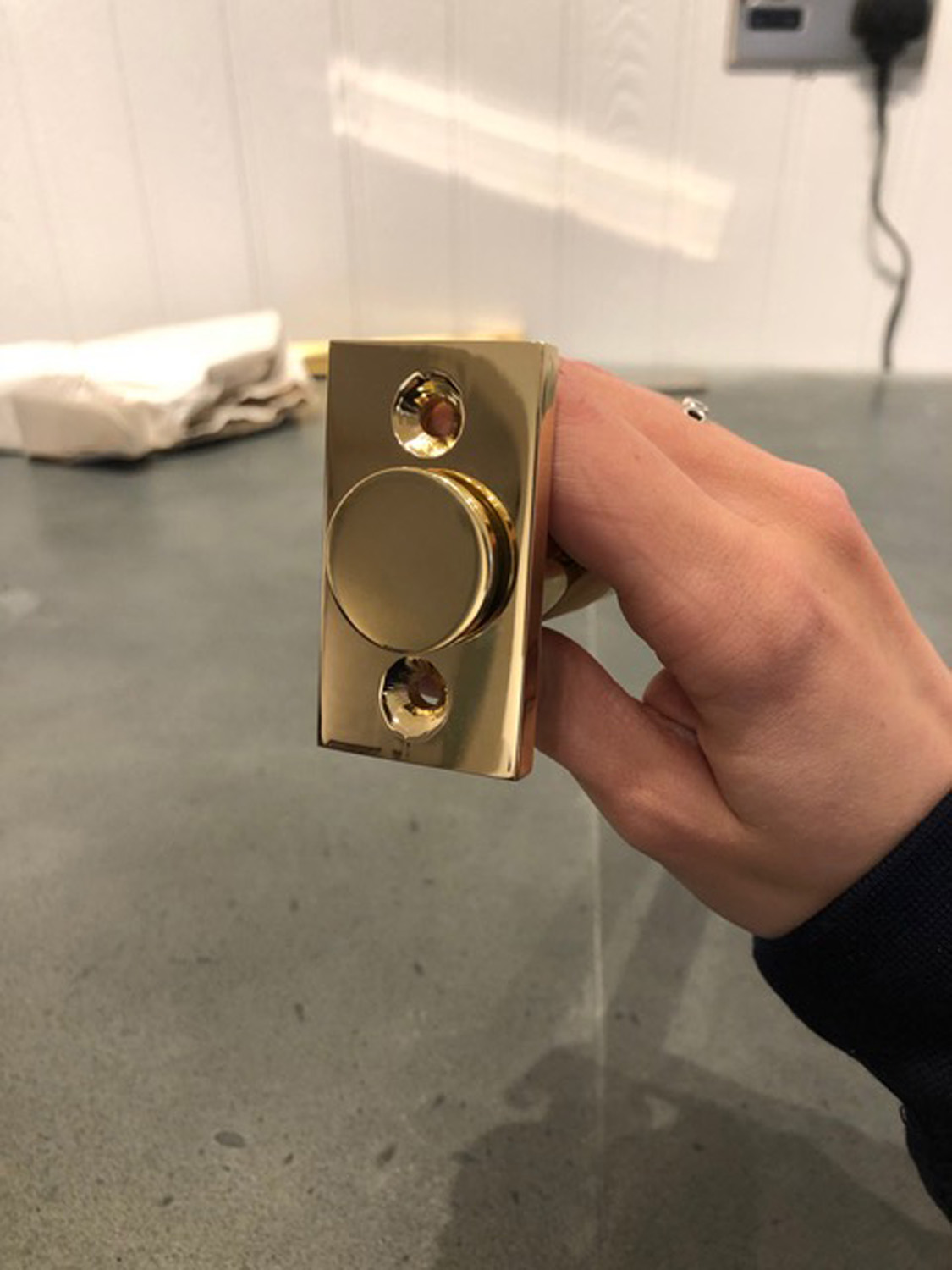A hand holding a shiny brass valet hook against a gray countertop background, with screws visible on the latch plate.