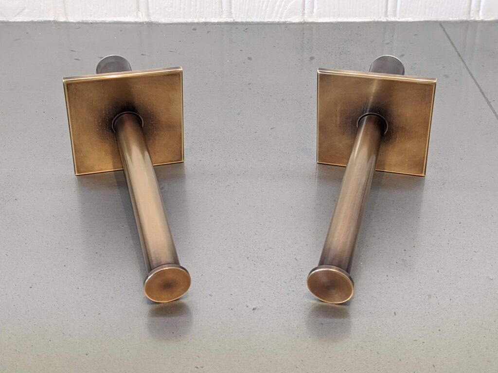 Two brass wall-mounted rods with square backplates are positioned parallel on a gray countertop, set against a white tiled wall.