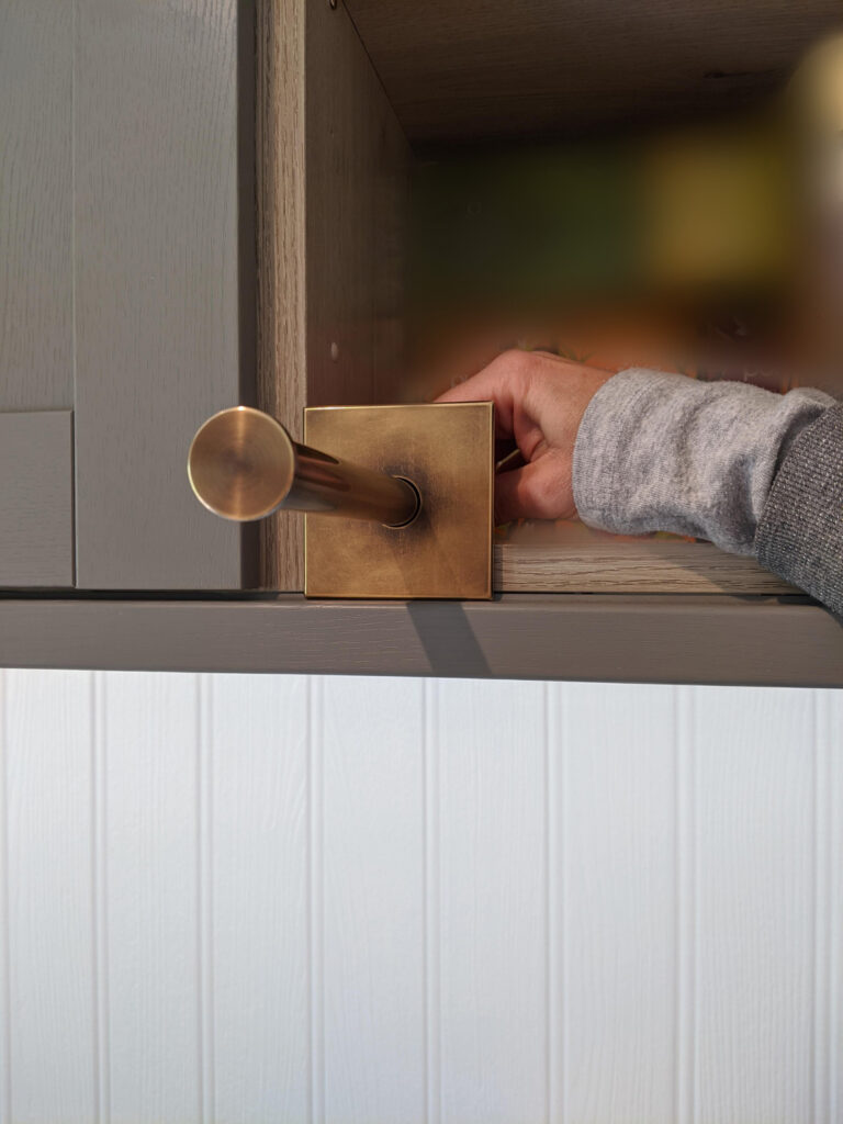 A person’s hand is holding a brass retractable valet hook to a cabinet, demonstrating the retracted state.