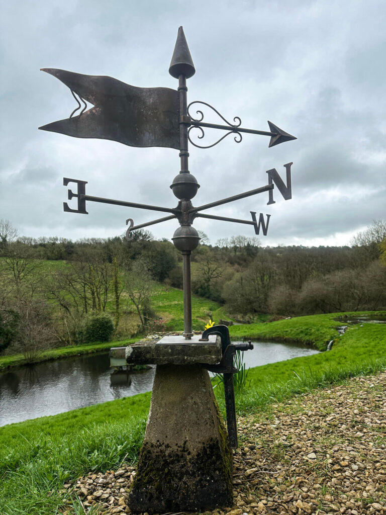 A weather vane showing cardinal directions stands on a stone base by a small pond, with grassy hills and trees in the background under a cloudy sky.