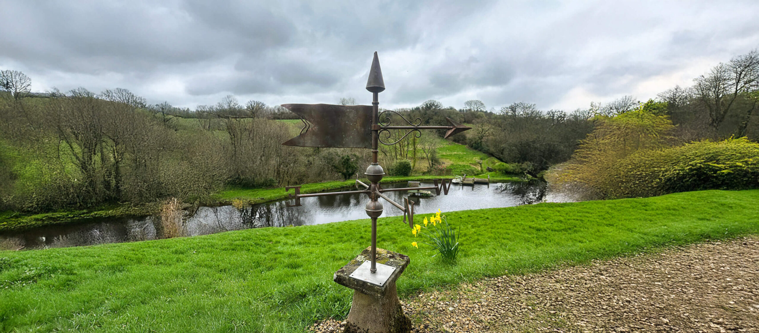 A weather vane stands on a grassy hill overlooking a river, with bare trees and cloudy skies in the background; clusters of yellow daffodils bloom near the weather vane.