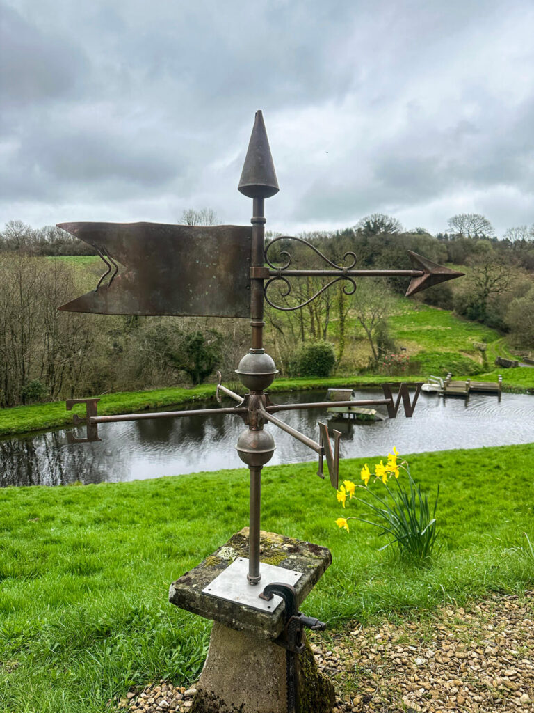 A metal weather vane with cardinal direction letters stands on a stone pedestal by a grassy area, overlooking a river and yellow daffodils, with trees and cloudy sky in the background.