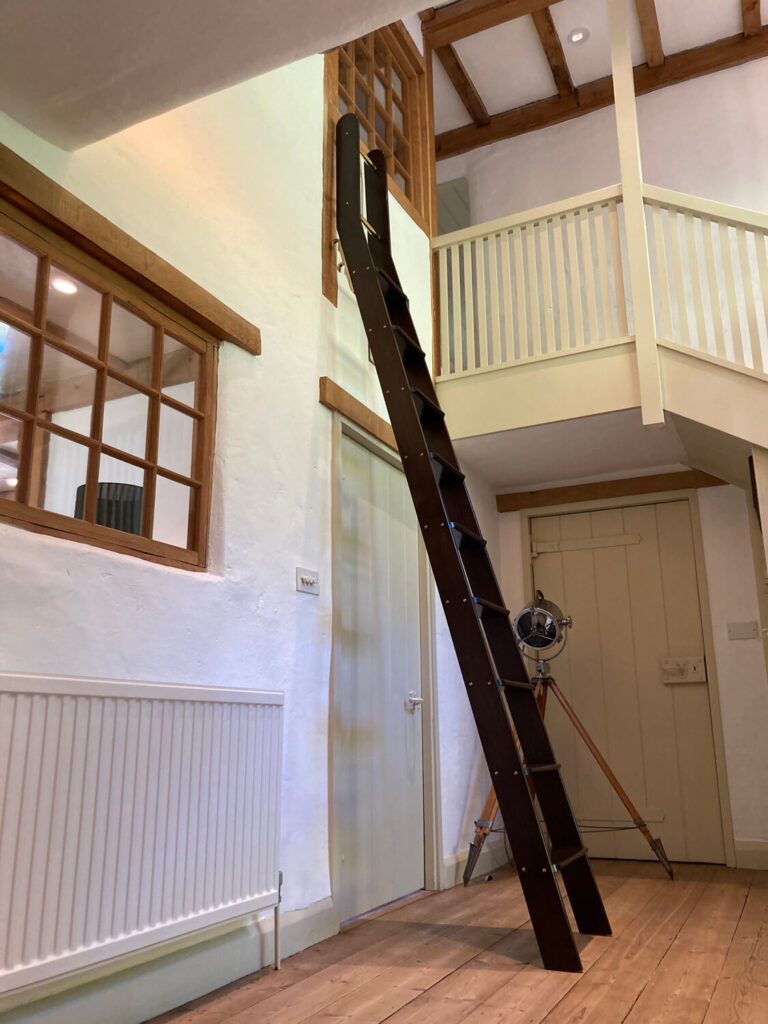 A dark wooden ladder leans against a high interior balcony in a room with white walls, wood trim, and wooden floors. There are windows, a radiator, and a tripod light nearby.