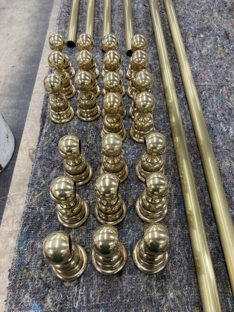 Rows of shiny brass posts and long brass poles are arranged neatly on a textured gray surface, likely prepared for assembly or installation.