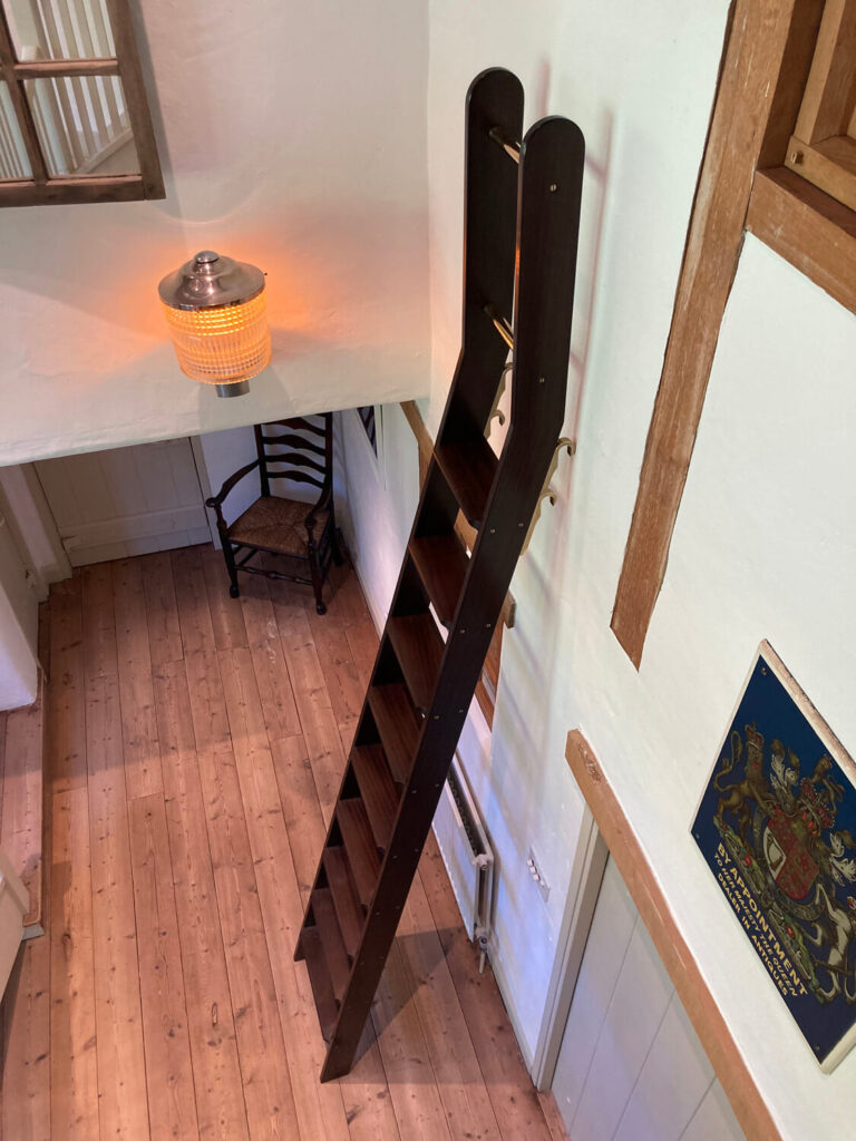 A dark wooden bookcase ladder attached to a tall white wall in a rustic room with wooden floors, a hanging lamp, a wooden chair, and a decorative wall plaque.