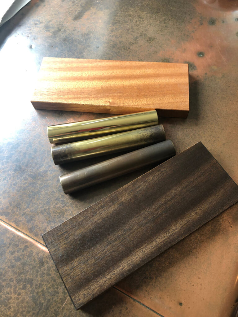 A photo shows two rectangular wooden blocks, one light and one dark, and three shiny metal rods with varying shades of brass, all placed on a reflective, mottled surface.