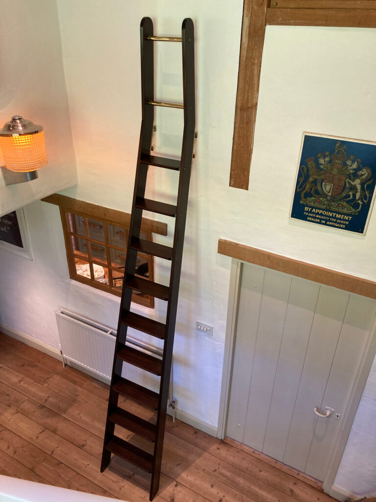 A cranked wooden ladder leans against a white wall, reaching up to a loft area. The room has wooden floors, a gray door, a window with wooden trim, a radiator, a wall lamp, and a framed art piece.