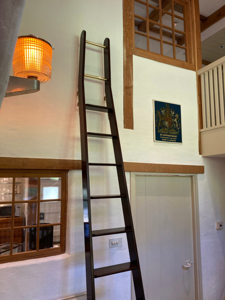 A tall, dark wooden ladder leans against a white wall inside a cozy room. There are wooden-framed windows, a glowing orange wall lamp, and a door below a sign with a crest and text.