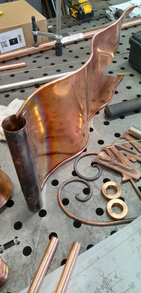 A shiny, curved copper flag is attached to a round copper tube on a perforated metal workbench, surrounded by copper rings, rods, scrolls, tools, and a sketch.