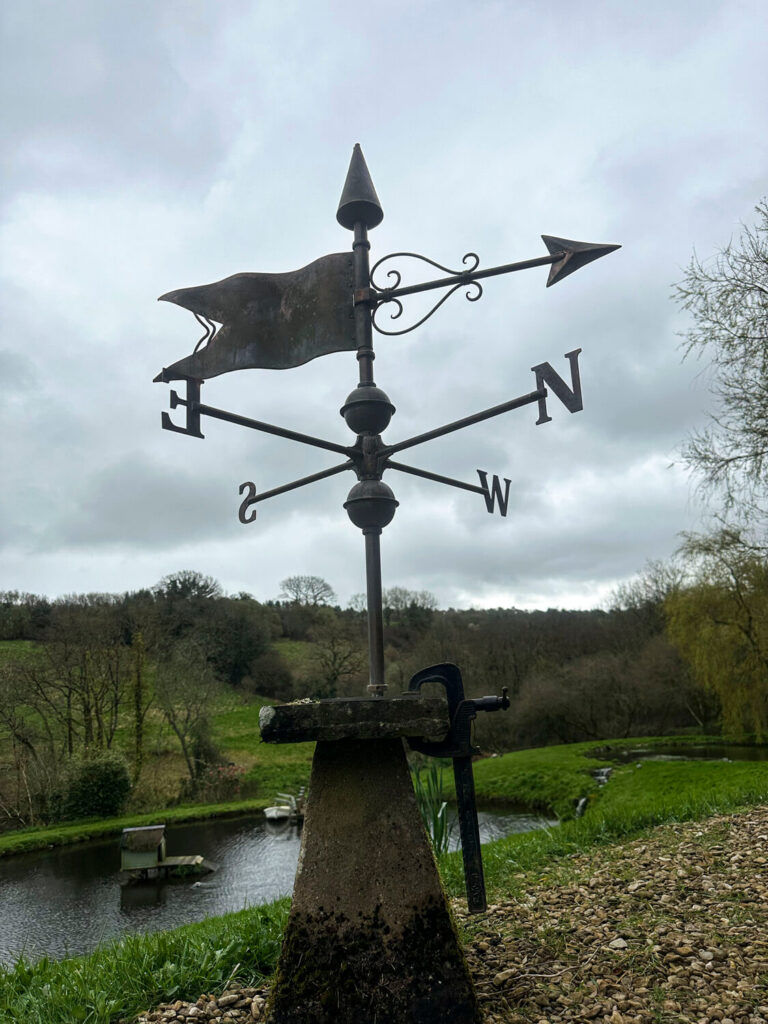 A metal weather vane with directional markers (N, S, E, W) stands on a stone base by a pond in a grassy landscape with trees and cloudy skies in the background.