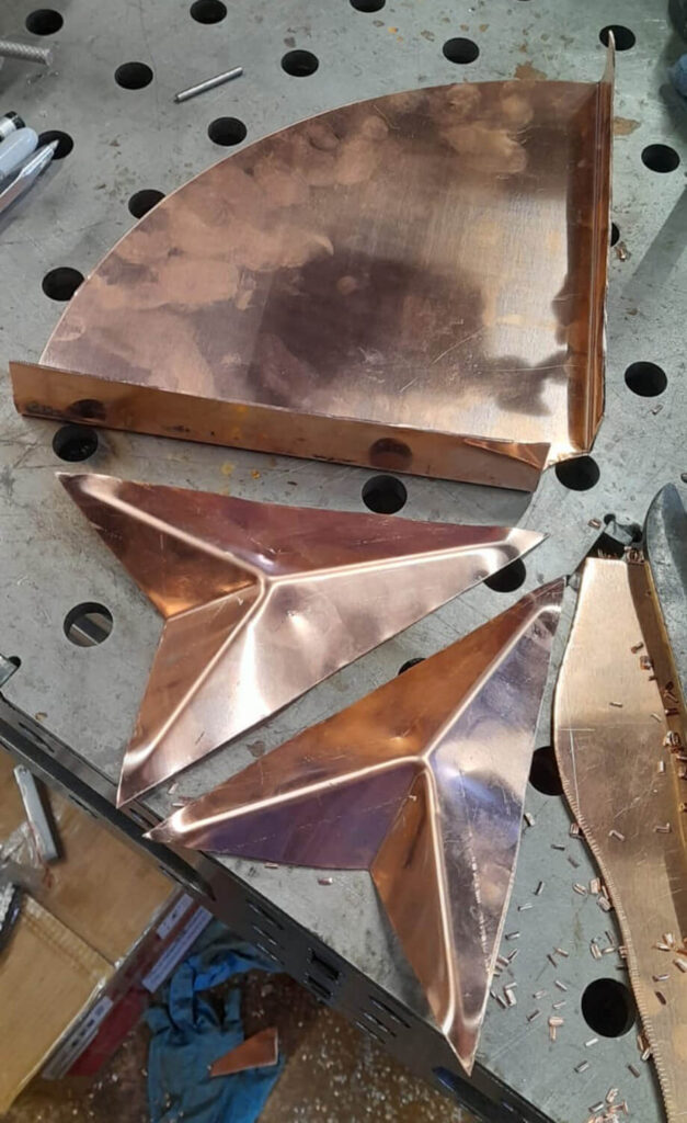 Three copper metal pieces rest on a perforated workbench: a large quarter-circle piece, and two triangular pieces with raised central folds. Metal shavings and tools are visible around the workspace.