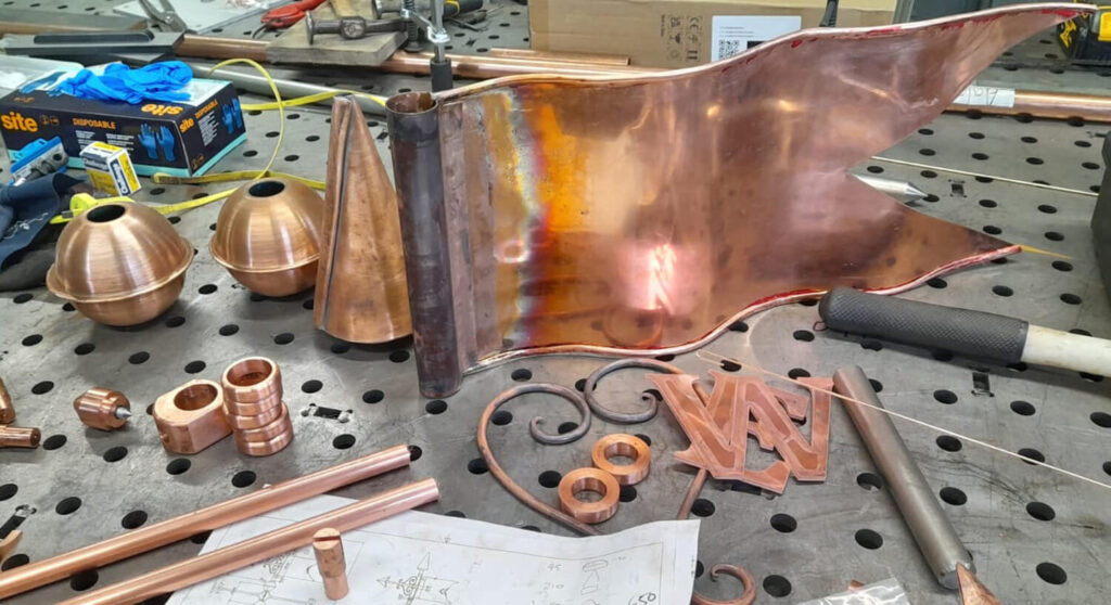 A workbench with various copper pieces, including a flag-shaped sheet, domes, rods, fittings, and decorative scrollwork, along with tools, papers, and boxes in the background.