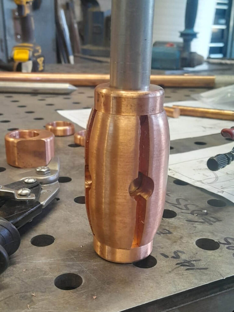 A polished copper mechanical part with precise grooves and cutouts is mounted on a metal rod, placed on a perforated workbench in a workshop setting with tools and diagrams visible in the background.