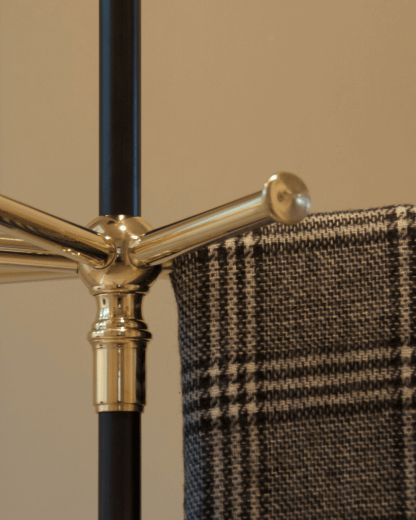 A close-up of a brass coat rack with arms extended, holding a black and white plaid scarf draped over one arm, against a neutral beige background.