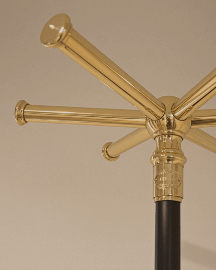Close-up of a shiny brass coat rack with several horizontal arms extending outward, mounted on a black pole, against a beige background.