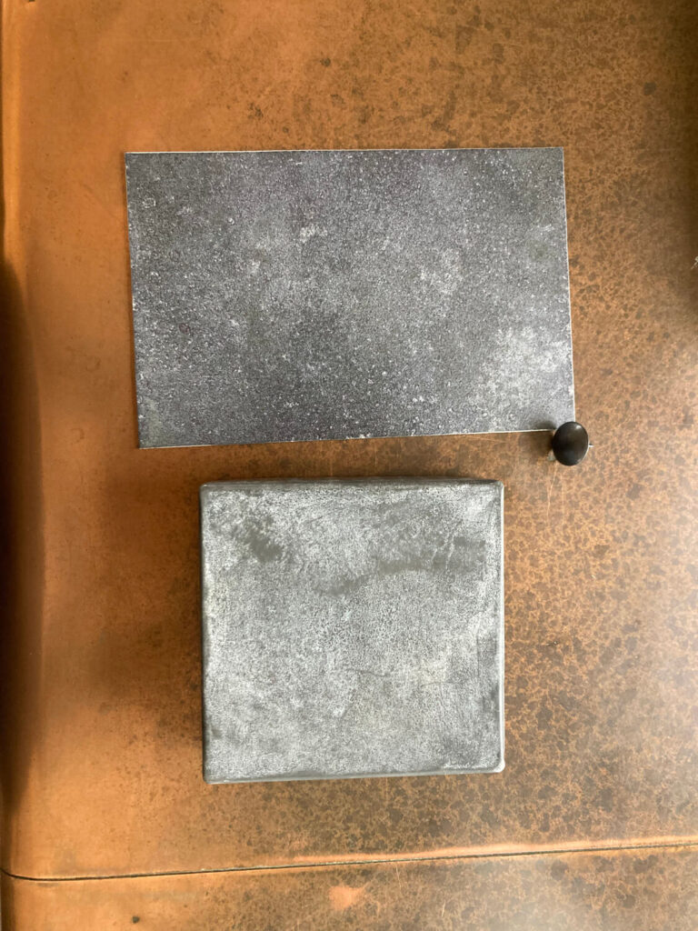 Two zinc metal samples that have been aged sit on a brass table; the upper plate is rectangular and has a small black knob attached to its right side, while the lower plate is square.