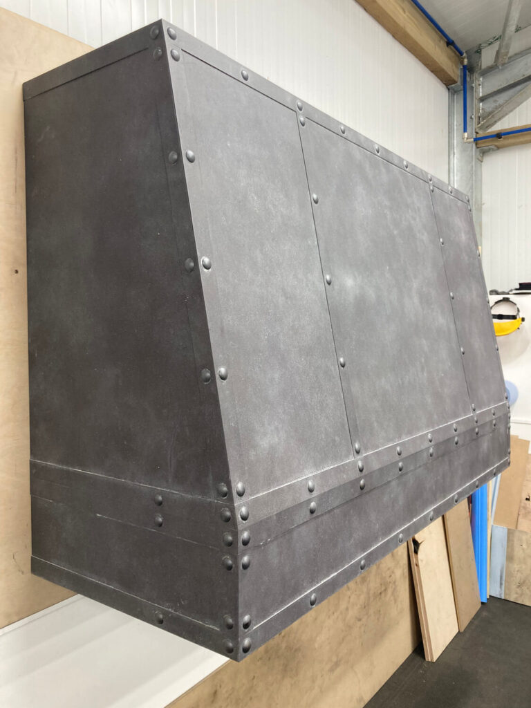 A large, industrial-style metal range hood with rivet details is mounted on a wall in a workshop or garage, surrounded by plywood boards and construction materials.