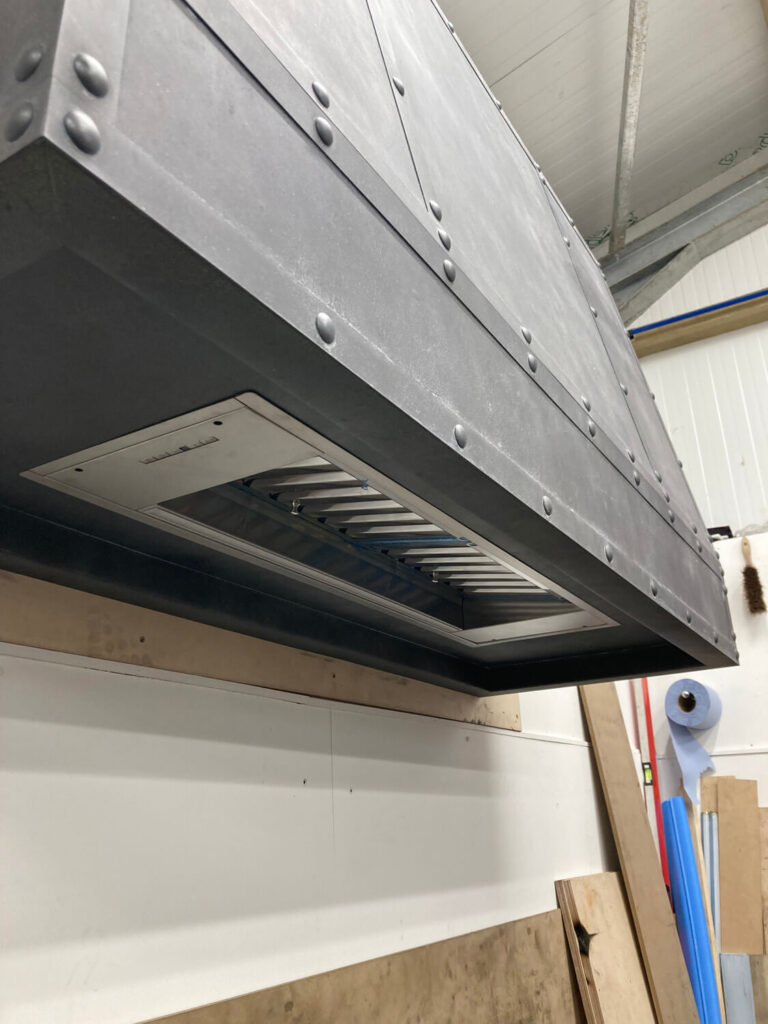 A large metal industrial kitchen hood with rivets is mounted above a wall in a workshop, with wooden boards and tools visible below and beside it.