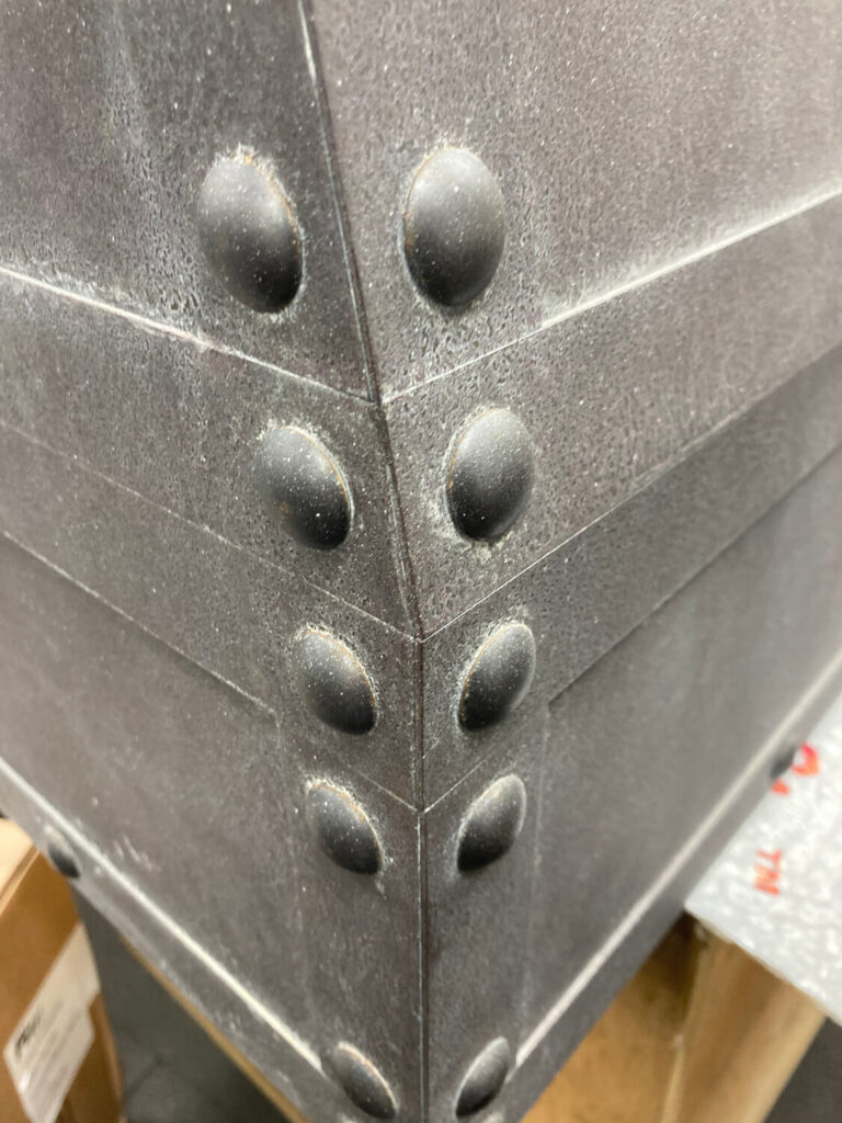 Close-up of the corner of bespoke cooker hood with rows of large, round rivets on both sides. Cardboard boxes and a textured surface are partially visible in the background.