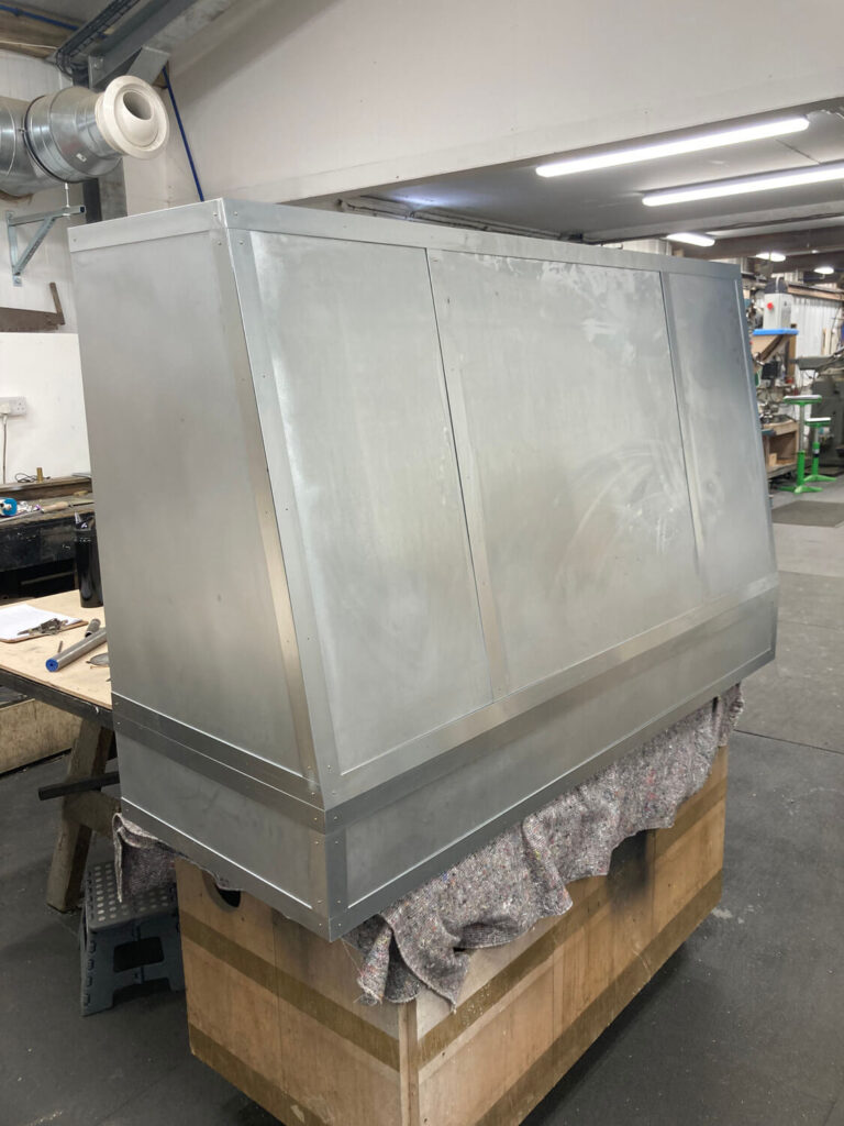 A large zinc cooker hood with angled panels is placed on a wooden crate in a workshop, surrounded by tools and industrial equipment.