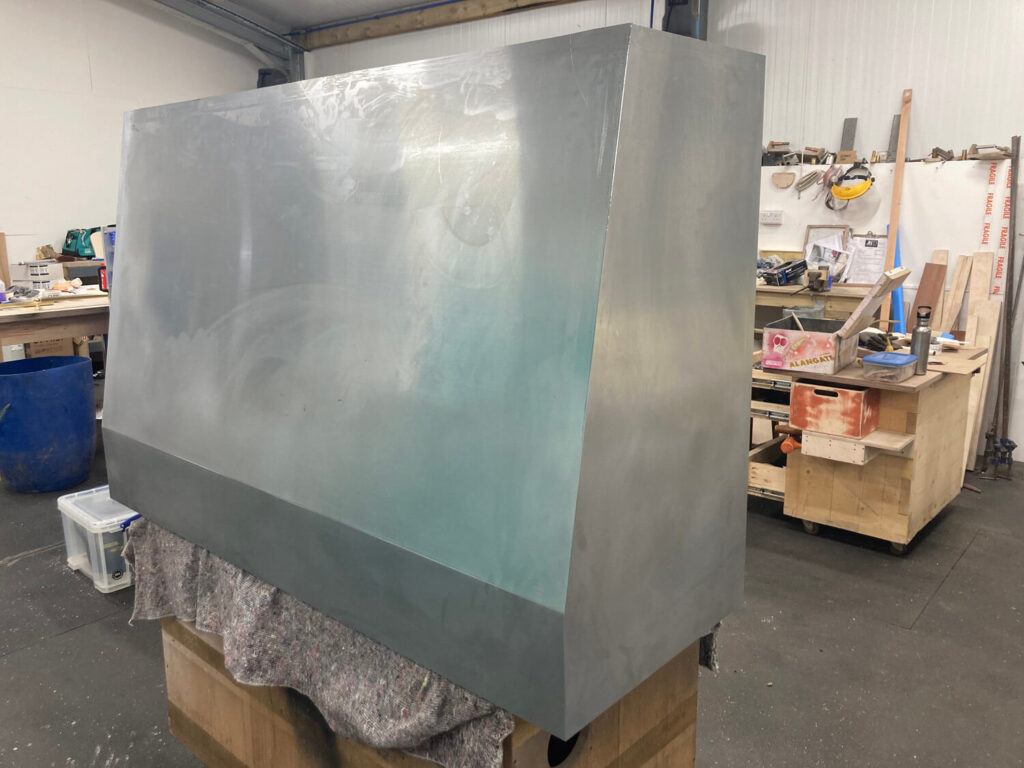 A large, industrial style handmade cooker hood with a slanted side sits on a covered wooden pallet in a workshop filled with tools, workbenches, and various materials.