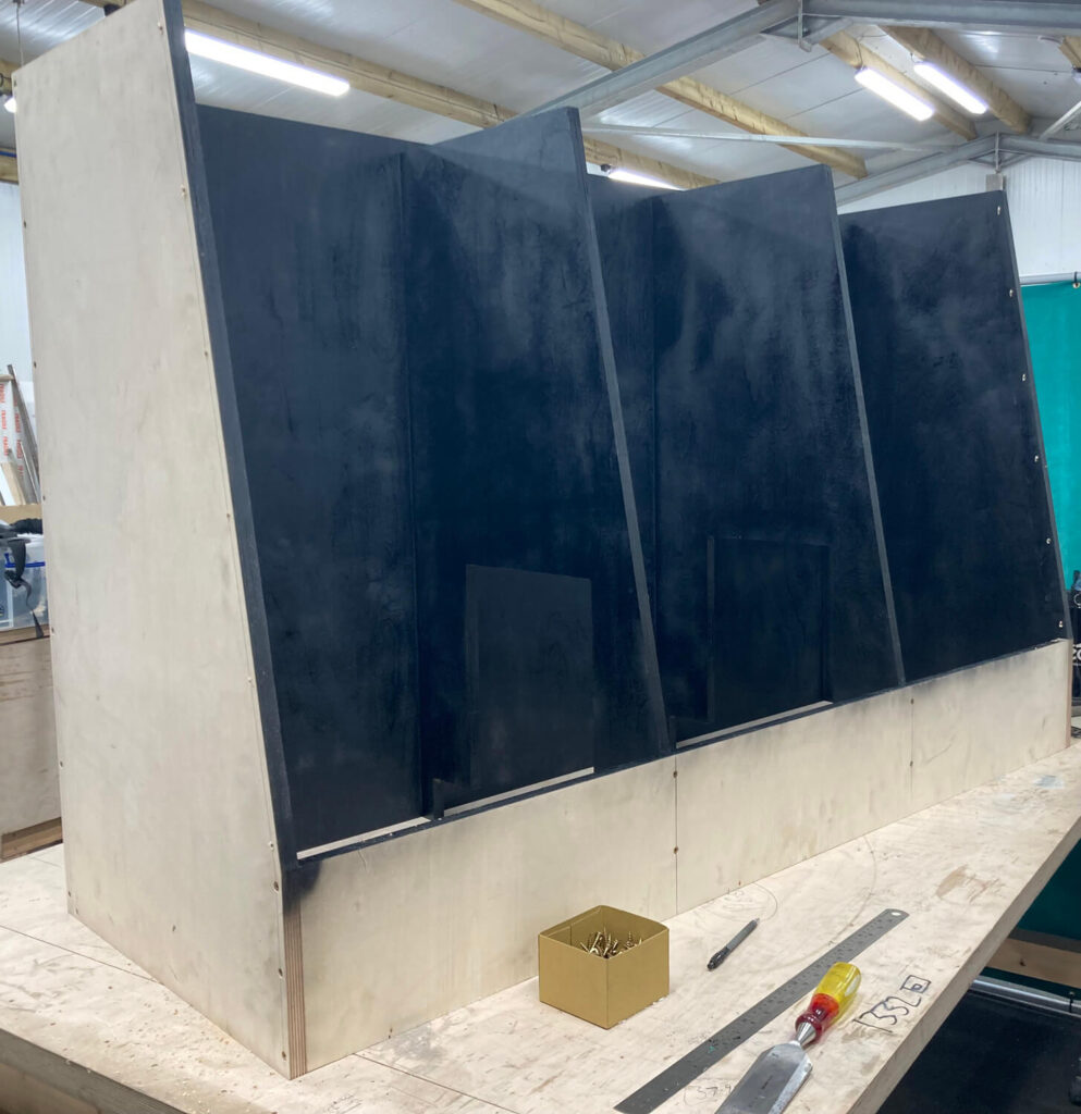A large wooden frame with three angled black panels stands on a workbench in a workshop. Tools and a small box of screws are on the table, with insulation visible on the ceiling above.