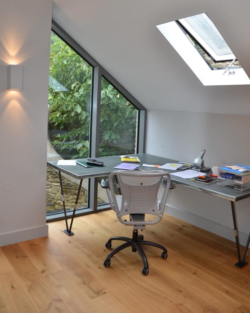 A modern home office with a zinc top desk, office chair, papers, books, and stationery. Large windows and a skylight let in natural light, with greenery visible outside. The room has light walls and wooden flooring.