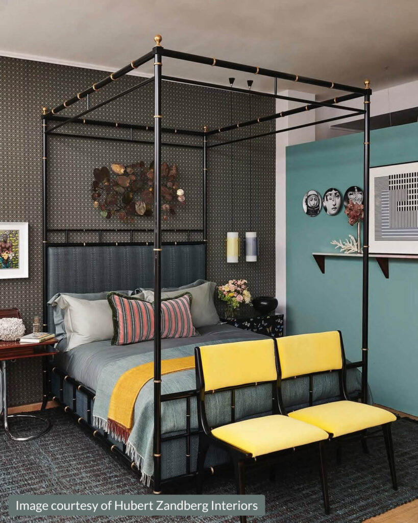 A modern bedroom with a black four-poster bed, colorful bedding, two yellow chairs at the foot, a dark patterned wall, eclectic decor, and a wall-mounted air conditioner above quirky wall art.