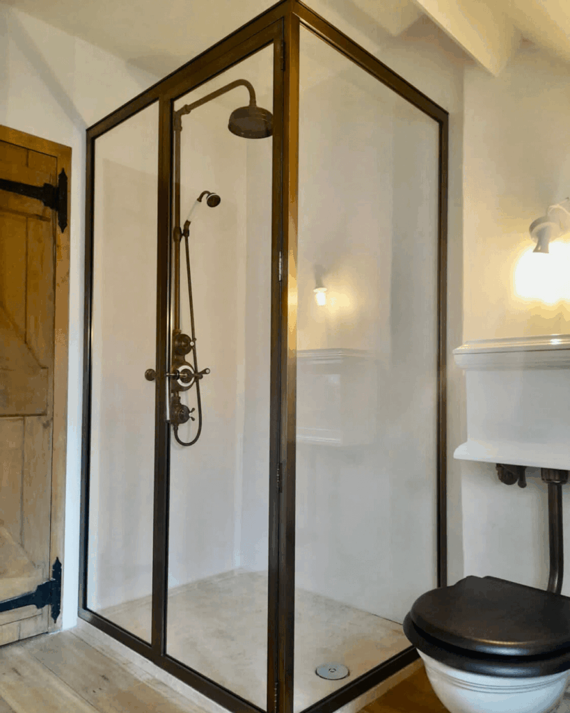 A modern glass shower enclosure with a dark metal frame and brass shower fixtures pairs with a rain showerhead, adjacent to a toilet with a dark seat and a wooden door in a bright bathroom.