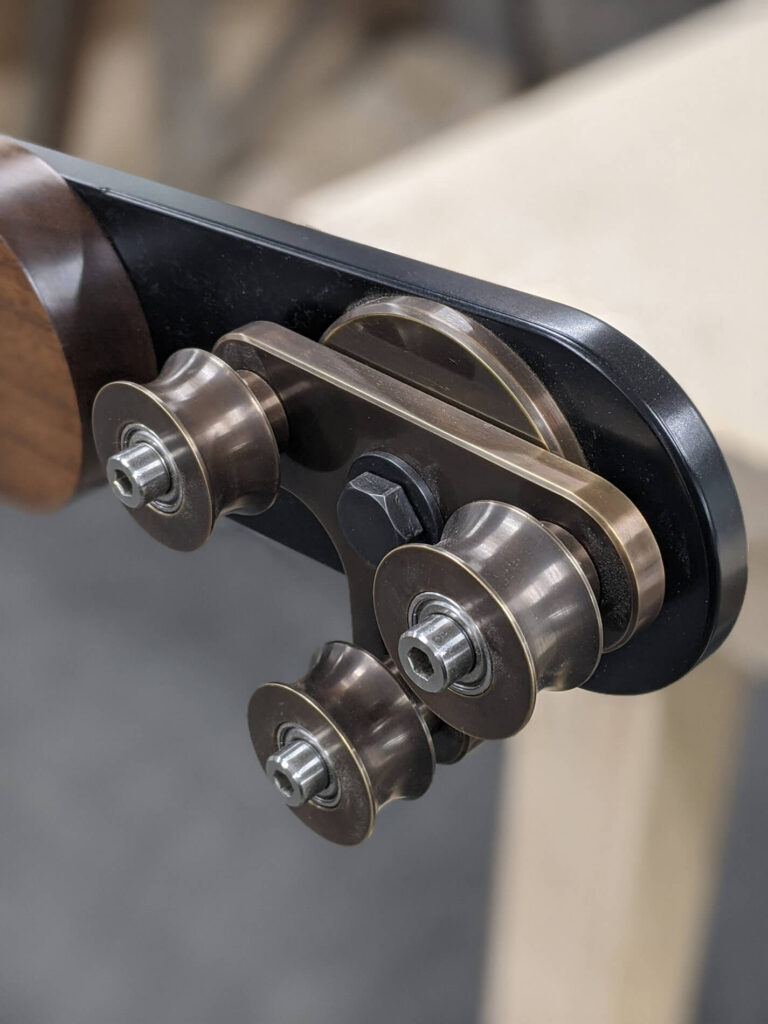 Close-up of a metal sliding door roller assembly with four bronze-coloured wheels attached to a black metal bracket, mounted on a wooden surface.