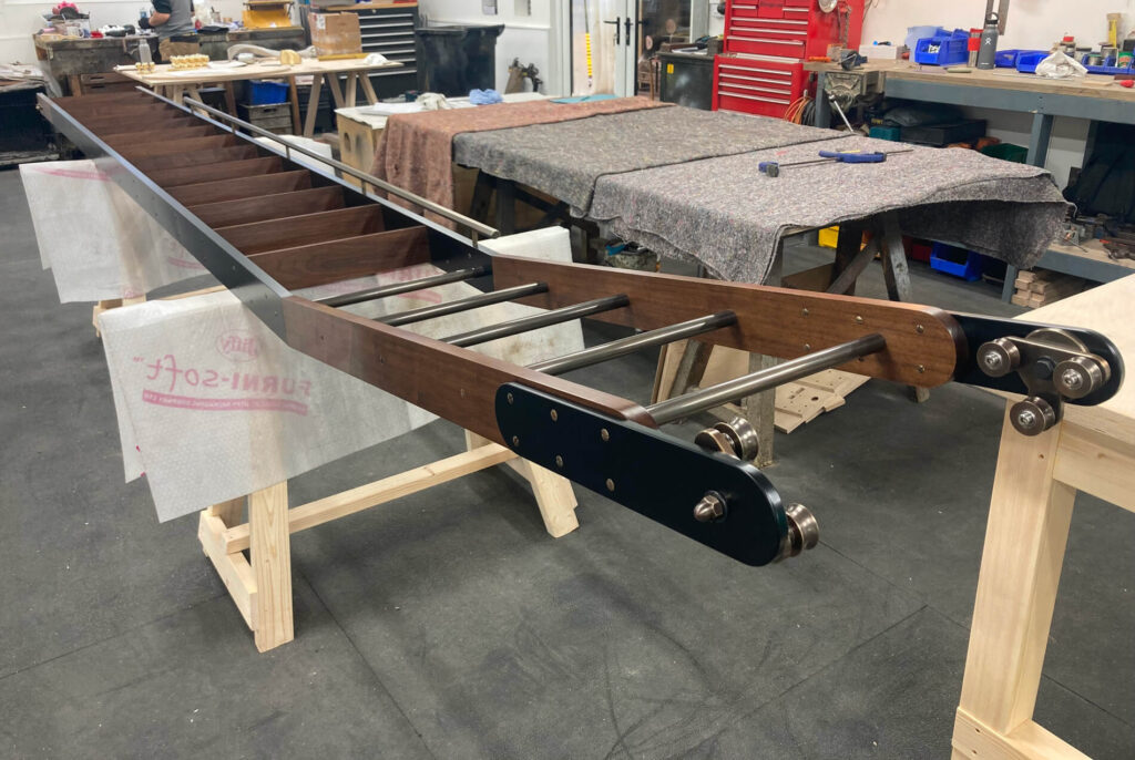 A large metal and wood ladder-like structure is supported by wooden saw-horses in a workshop, with various tools, equipment, and workbenches visible in the background.