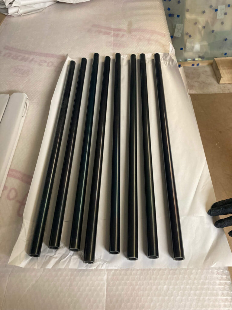 Eight black cylindrical metal pipes are neatly arranged in parallel on a sheet of white protective material, with part of the work area and other packaging visible around them.