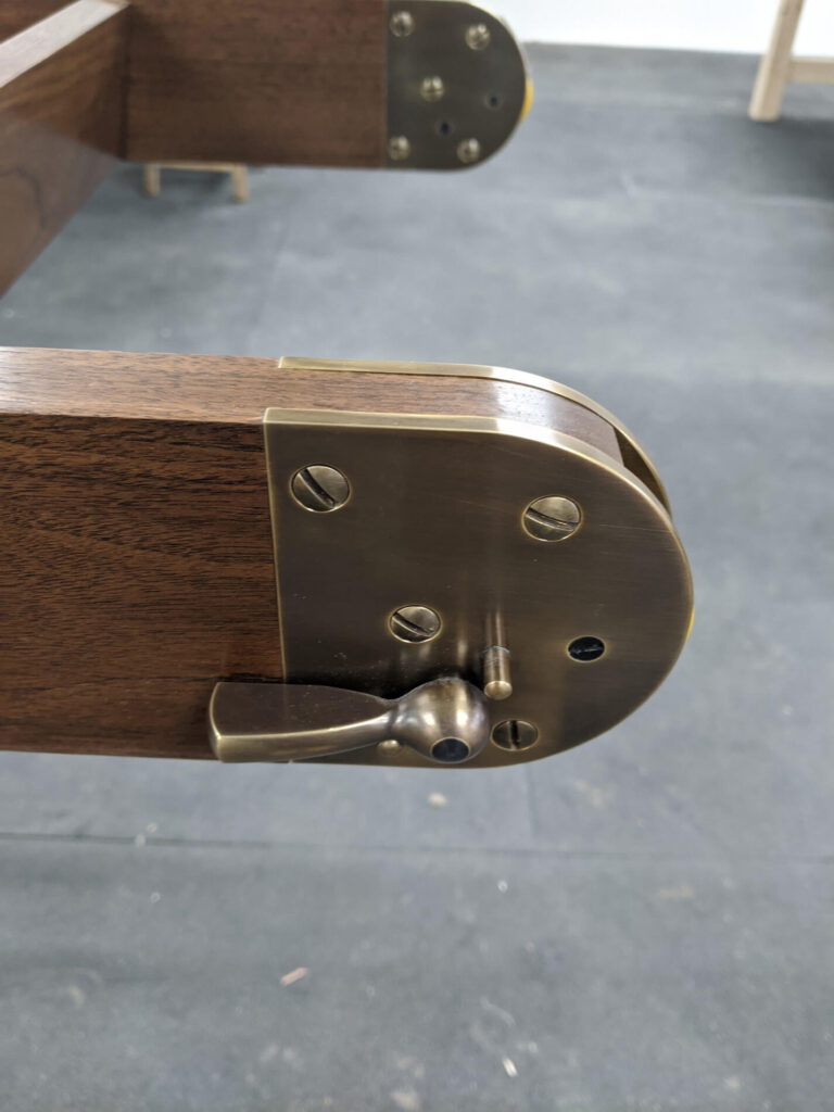 Close-up of a brass mechanical latch attached to the end of a dark wooden beam, with visible screws and lever handle, on a dark grey floor background.